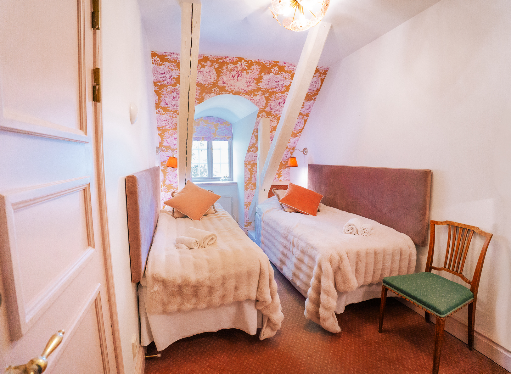 Tvåbäddsrum med två enkelsängar, rosa sammetsgavlar och orange kuddar mot en orange toile-tapet. Välvt fönster i bakgrunden. Ulvhäll Herrgård, Strängnäs.