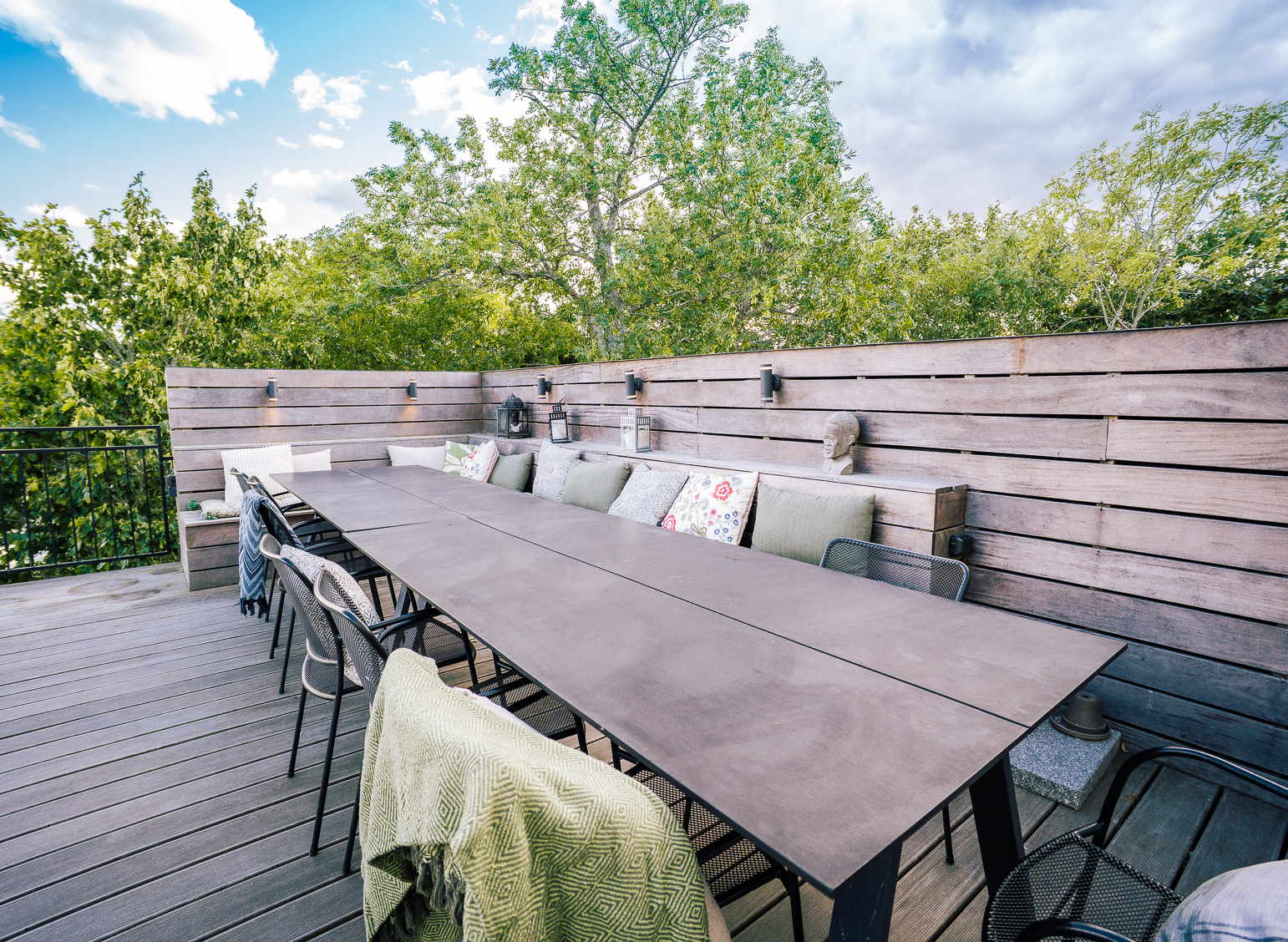 Privat terrass tillhörande sviten, med långt matbord, svarta stolar och inbyggd bänk med färgglada kuddar längs träpanelvägg. Omgiven av lummiga träd mot blå himmel. Ulvhäll Herrgård, Strängnäs.